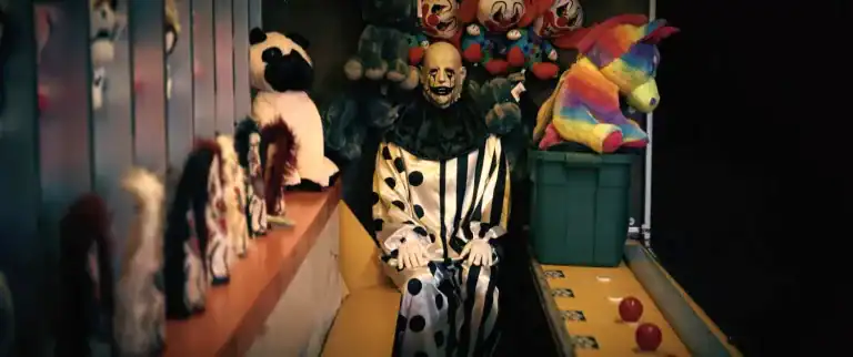 A sinister clown figure dressed in a striped costume sits among carnival prizes in Hell House LLC Lineage.