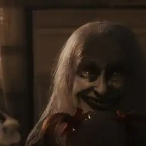 A sinister ghostly figure with pale skin and a wide smile appears in a dark room, holding a doll, from The Conjuring: Last Rites.