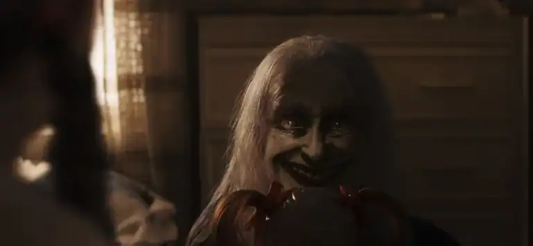 A sinister ghostly figure with pale skin and a wide smile appears in a dark room, holding a doll, from The Conjuring: Last Rites.