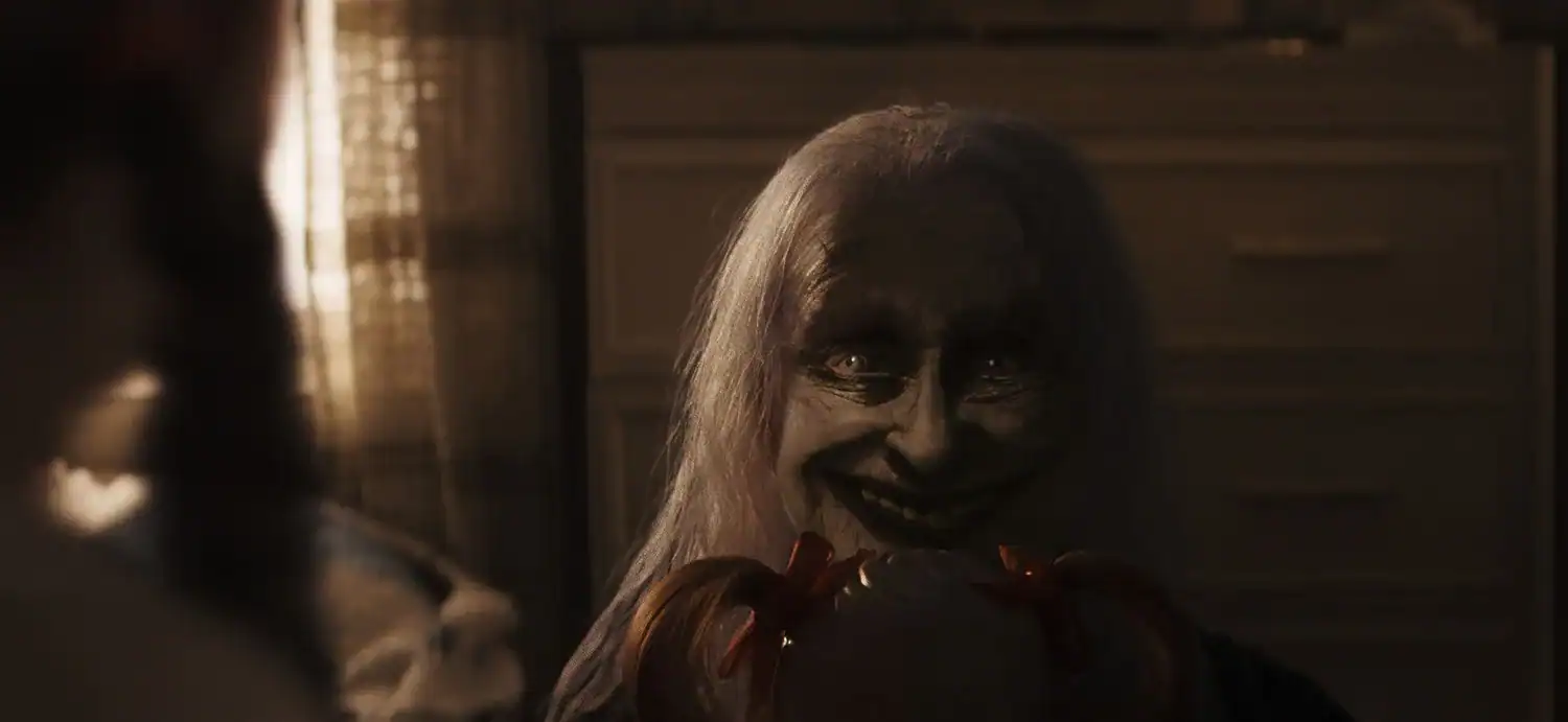A sinister ghostly figure with pale skin and a wide smile appears in a dark room, holding a doll, from The Conjuring: Last Rites.