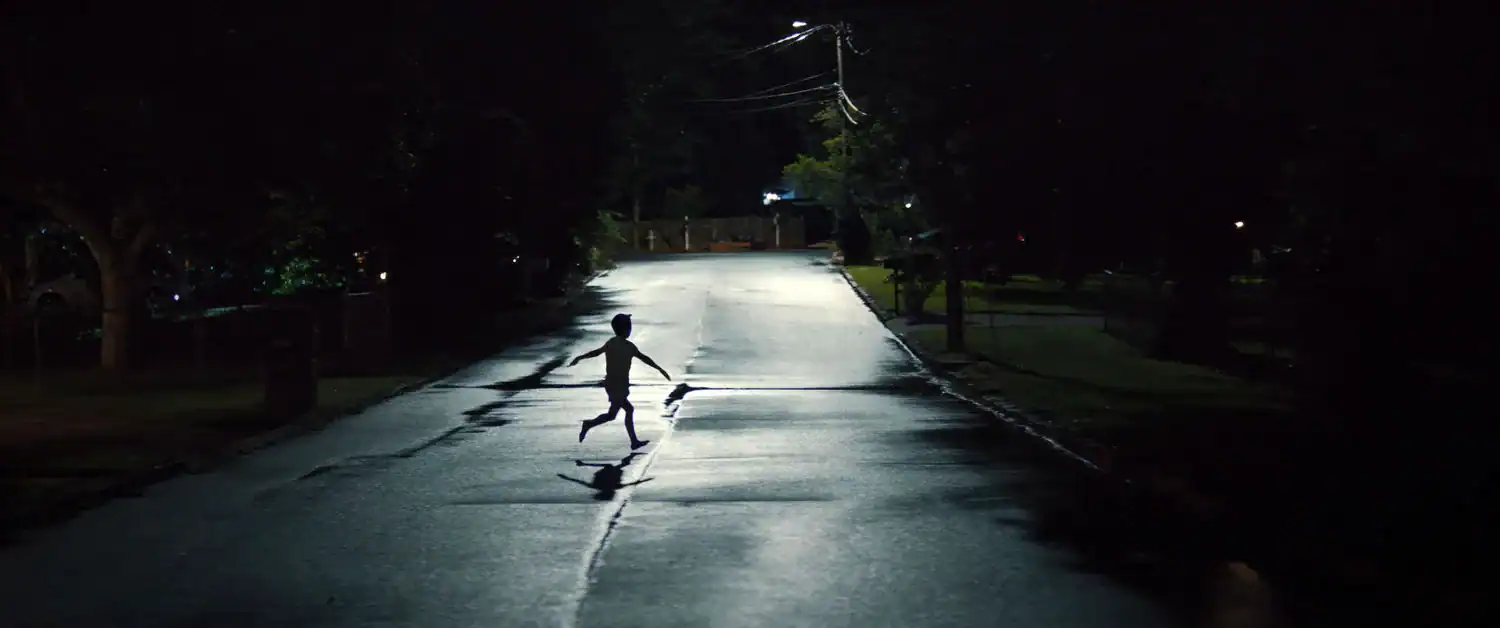 A child runs across a dark suburban street at night in a suspenseful scene from the movie Weapons (2025).